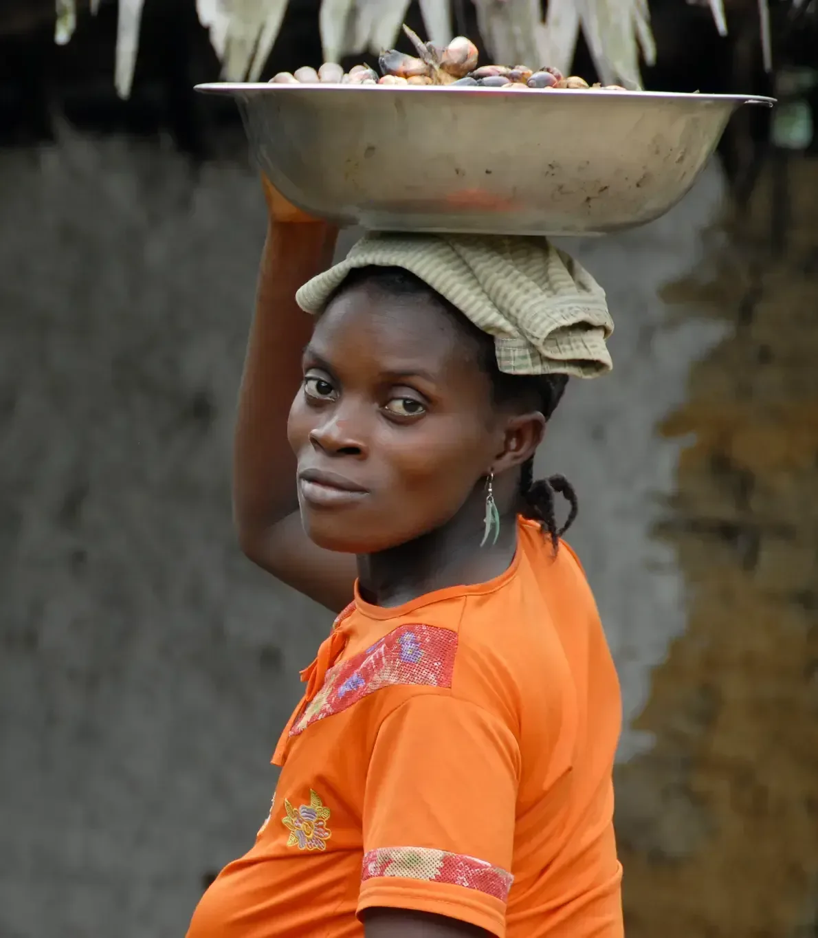 A woman in an orange shirt balances a metal bowl on her head.