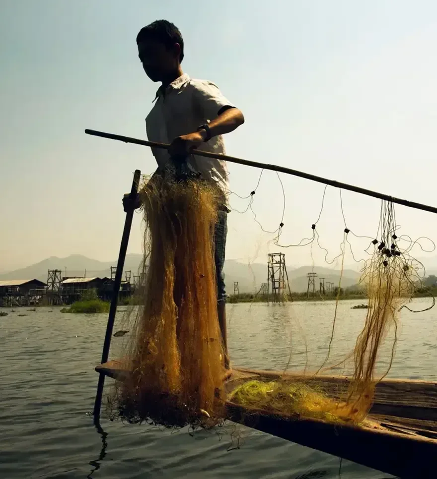 A fisherman balancing on his boat while preparing a large net on a calm lake.